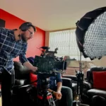 An image of a white male camera operator with a beard standing over his camera. There is lighting equipment around him. He is preparing to shoot an interview for a charity. The director is behind him. You can't see him but trust me, he's really handsome.