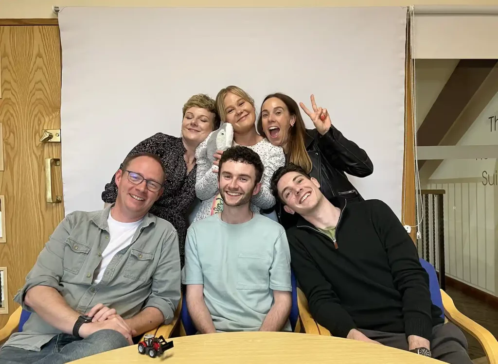A photo of Jess' last team day. The full team of six people are behind a table, posing with Jess in the middle. The image shows 6 people smiling. There's a toy tractor on the table in front because of the amount of agriculture video production the company gets involved in.