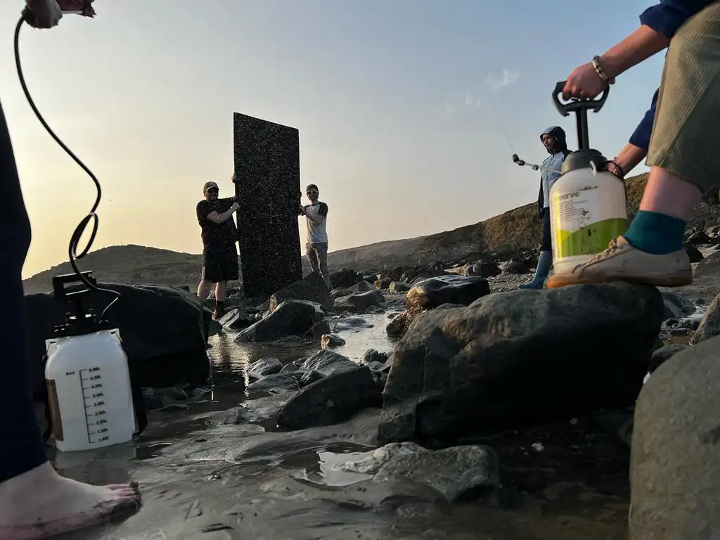 An image taken from the video shoot being referenced in the blog post. An actress is in a waterproof jacket avoiding the rain. But it's very sunny, so the crew have gathered a number of handheld water sprayers and are replicating rain. Two men are holding a large black fabric in a frame to block the direct sun on the actress.