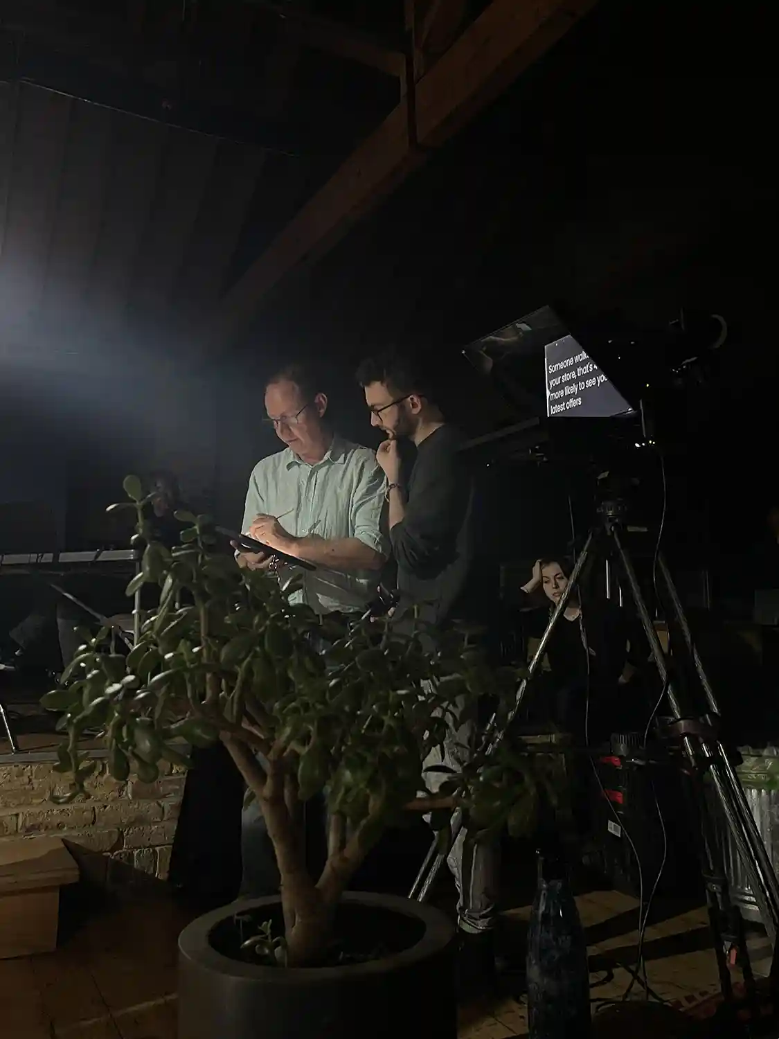 Image from the set of a startup video. Features 2 men standing in a dark room looking at an iPad. There is an autocue in the foreground with text visible.