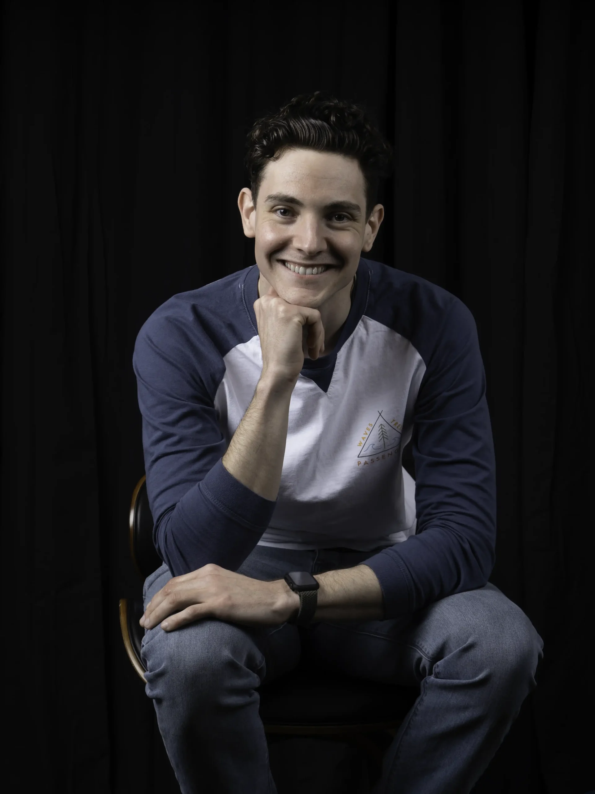 A portrait style photo of a man sitting on a stool. He's one of the video production team and is set against a black background. He's leaning forwards with one hand resting under his chin. he's smiling.