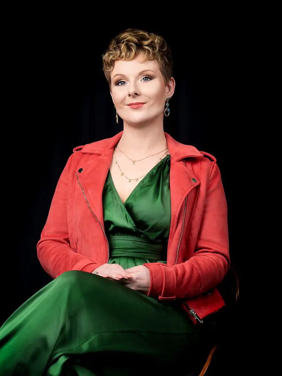 A portrait style photo of a woman against a black background. She's the creative producer for Vermillion Films and is wearing a shiny green dress trouser pant suit thing with a light red suede jacket.