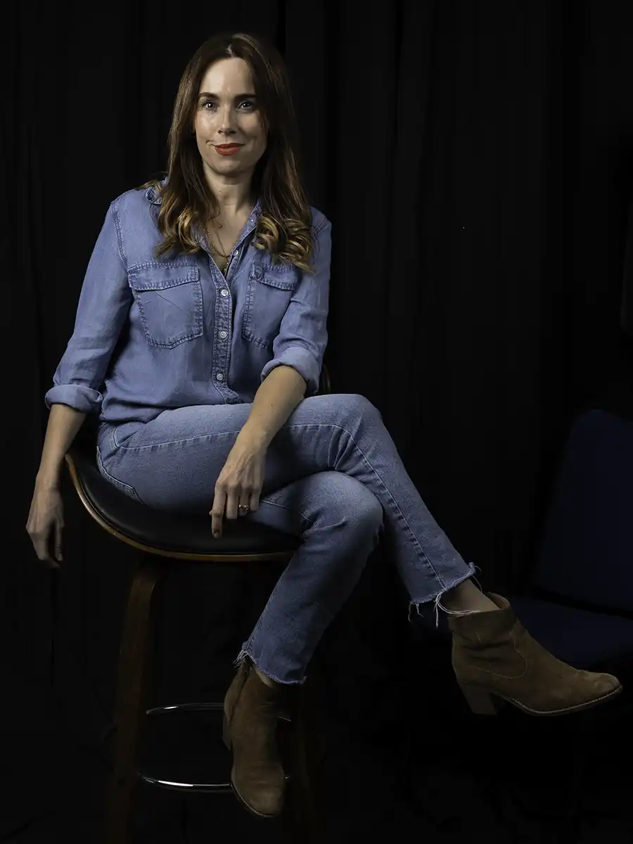 A portrait style photo of a woman from our video production team seated on a stool. She's wearing double denim.